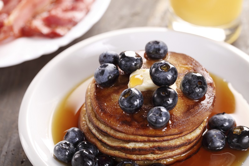 Stack of pancakes with blueberries, plate with bacon and glass with orange juice.